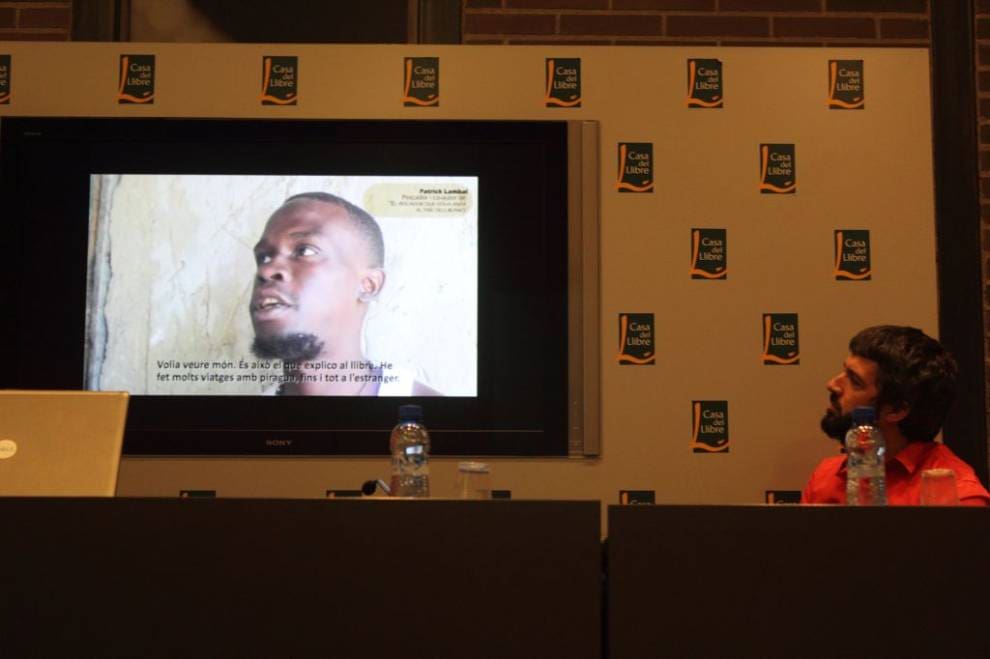 Jordi Tomàs observa un vídeo en el Patrick Lambal explica desde Senegal porqué ha participado en el libro. Ambos firman "El pescador que volia anar al país dels blancs".